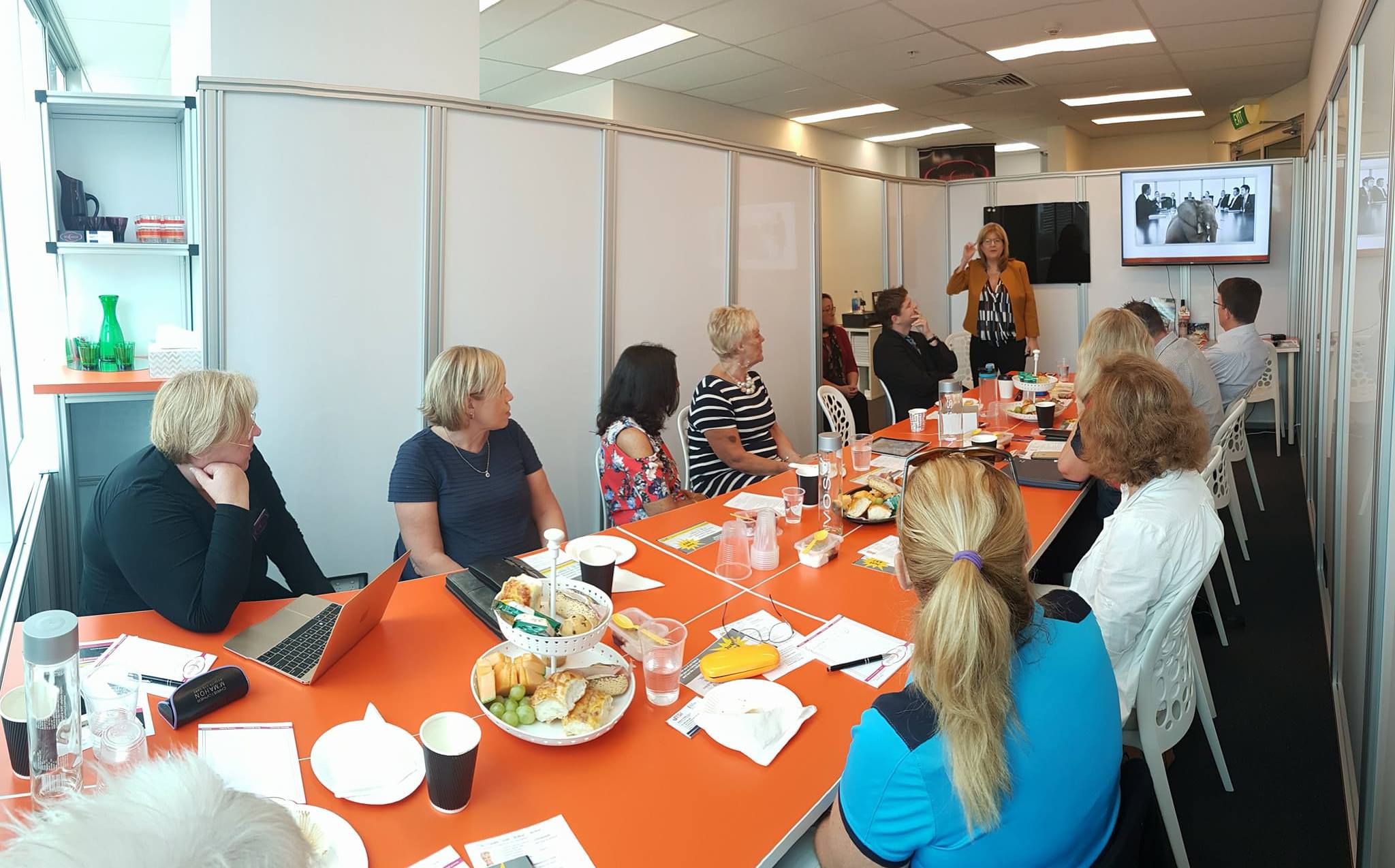 an image of sales and brand authority coach, Janeen Vosper running training for a group of business leaders in a room all sitting each side of a long orange table